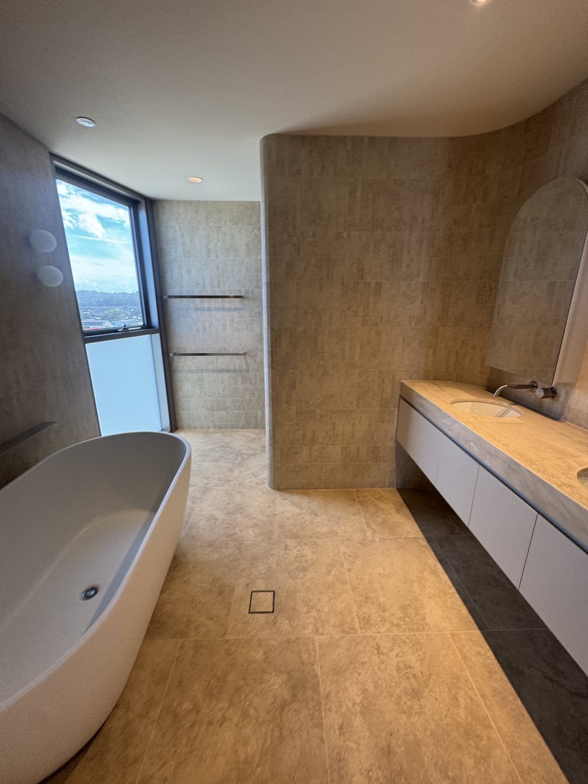 Freestanding bath beside a full-height window with curved travertine walls in a Gold Coast bathroom renovation
